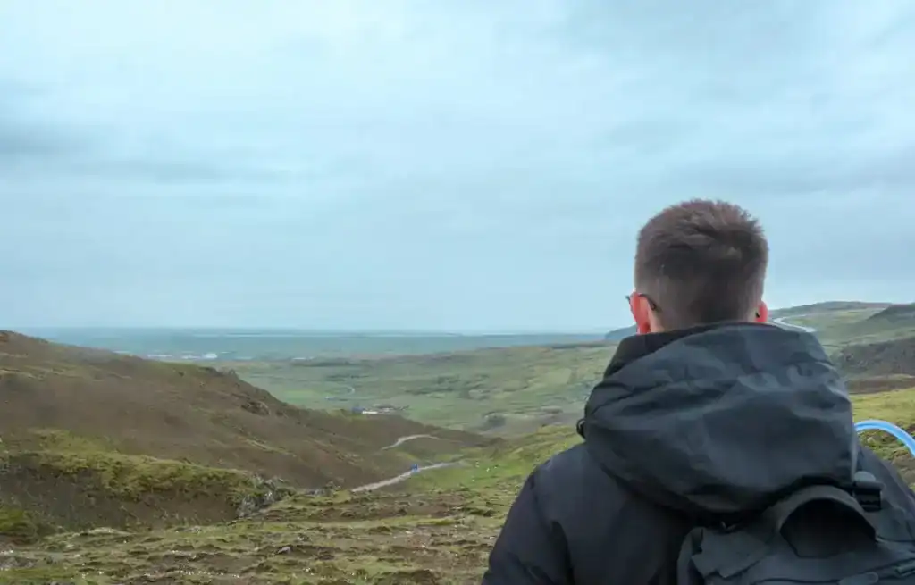 Back view of a man in a black jacket overlooking a green valley and distant coastline under cloudy sky.