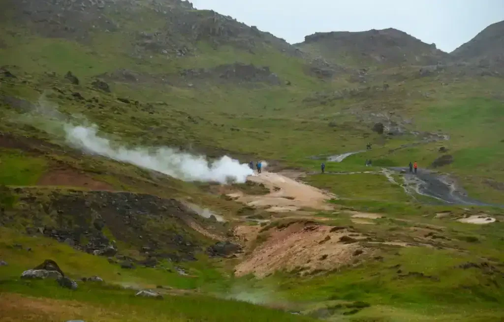 People walking near steaming geothermal area in a green mountainous landscape in Iceland.