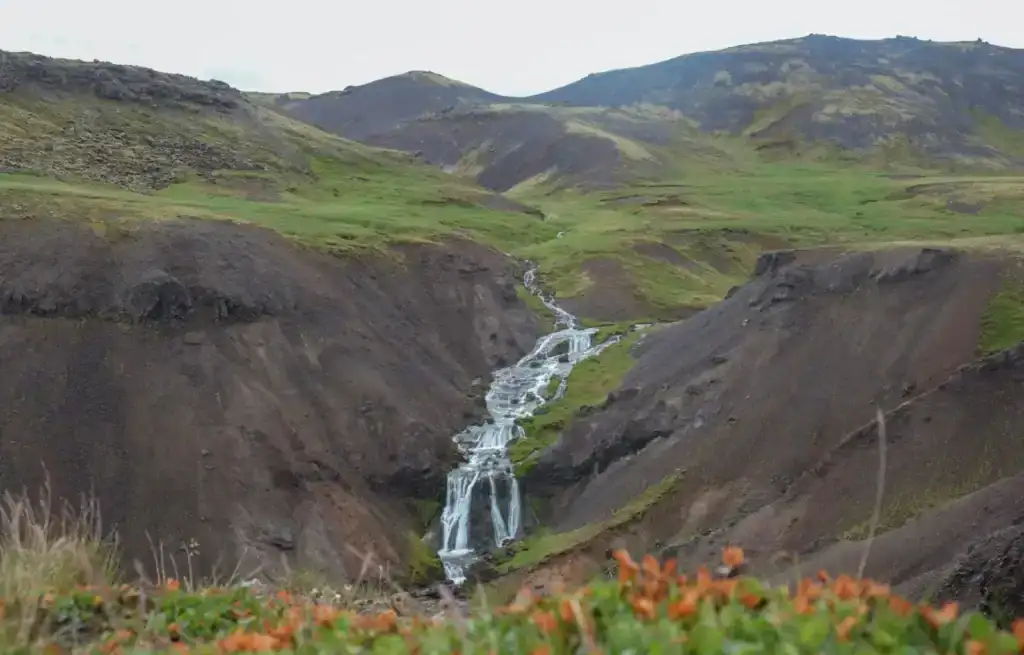 Small waterfall flowing through a rugged green valley with hills in the background.