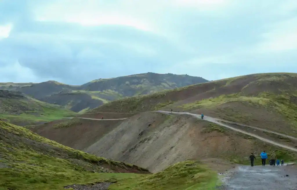 Hikers walking along a trail in a green, hilly landscape with volcanic terrain and distant mountains under a cloudy sky.