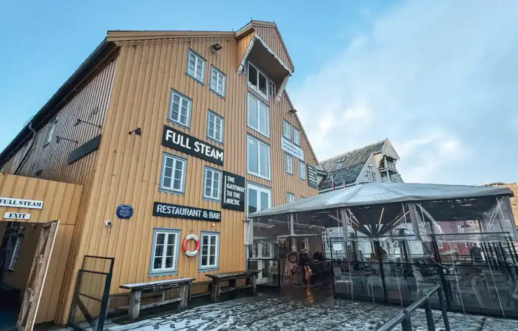 Wooden building with signs for Full Steam restaurant and bar, with outdoor seating area and glass enclosure, under a cloudy,.