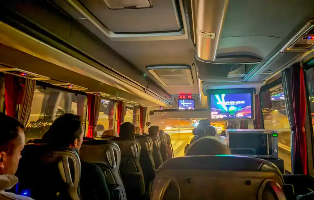 Passengers watching a screen on a bus at night with city lights outside.