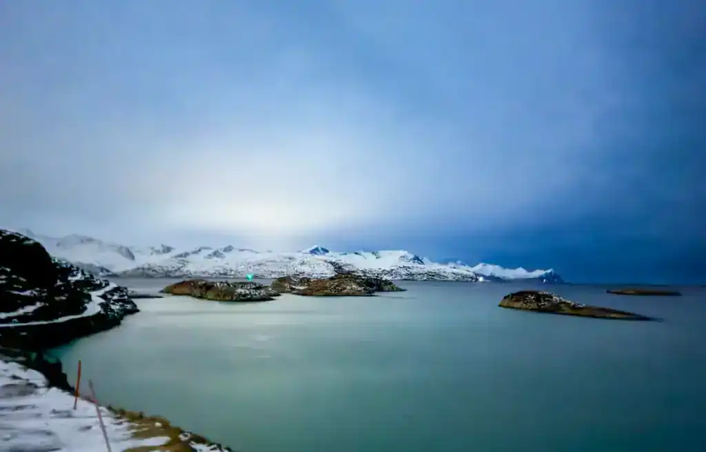 Snow-covered mountains and islands seen across a calm, icy fjord under a cloudy sky.