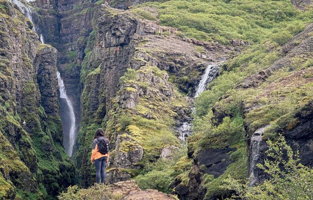 Hiker standing near waterfalls in a lush green canyon with moss-covered rocks.