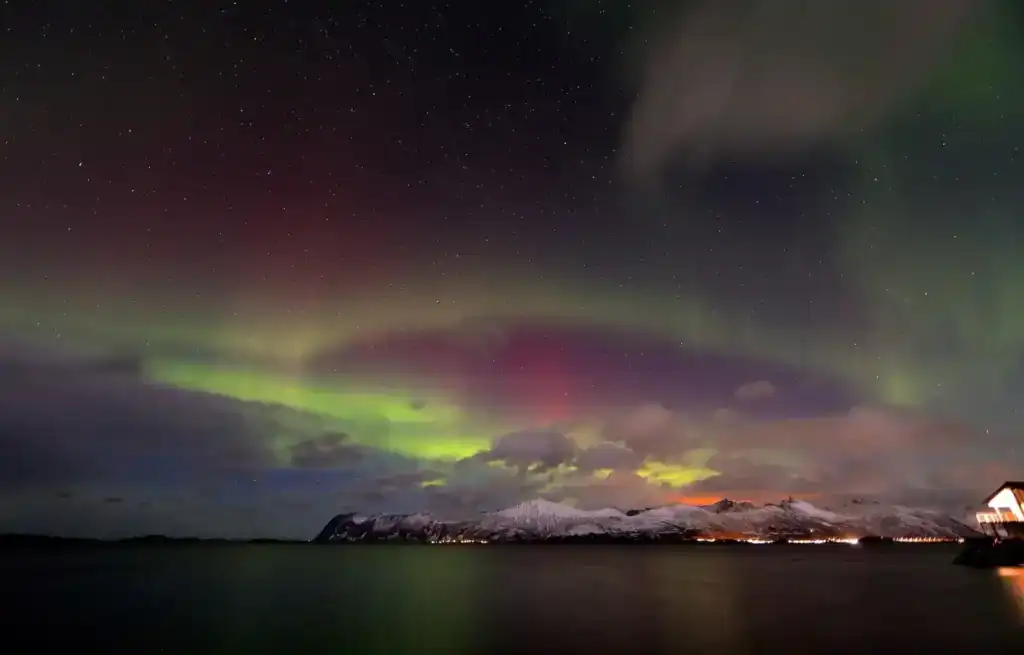 Northern Lights dance over snow-capped mountains and a dark fjord in Tromso, Norway, under a starry sky.