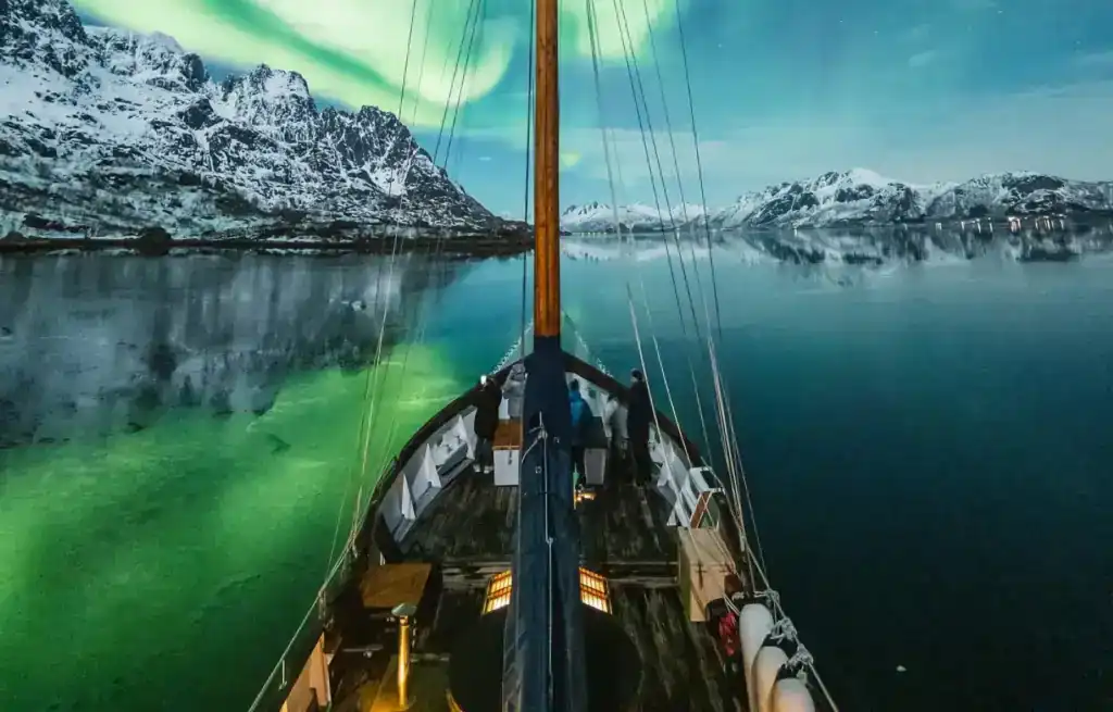 View from a boat sailing through calm waters with snow-capped mountains and the Northern Lights in the sky.
