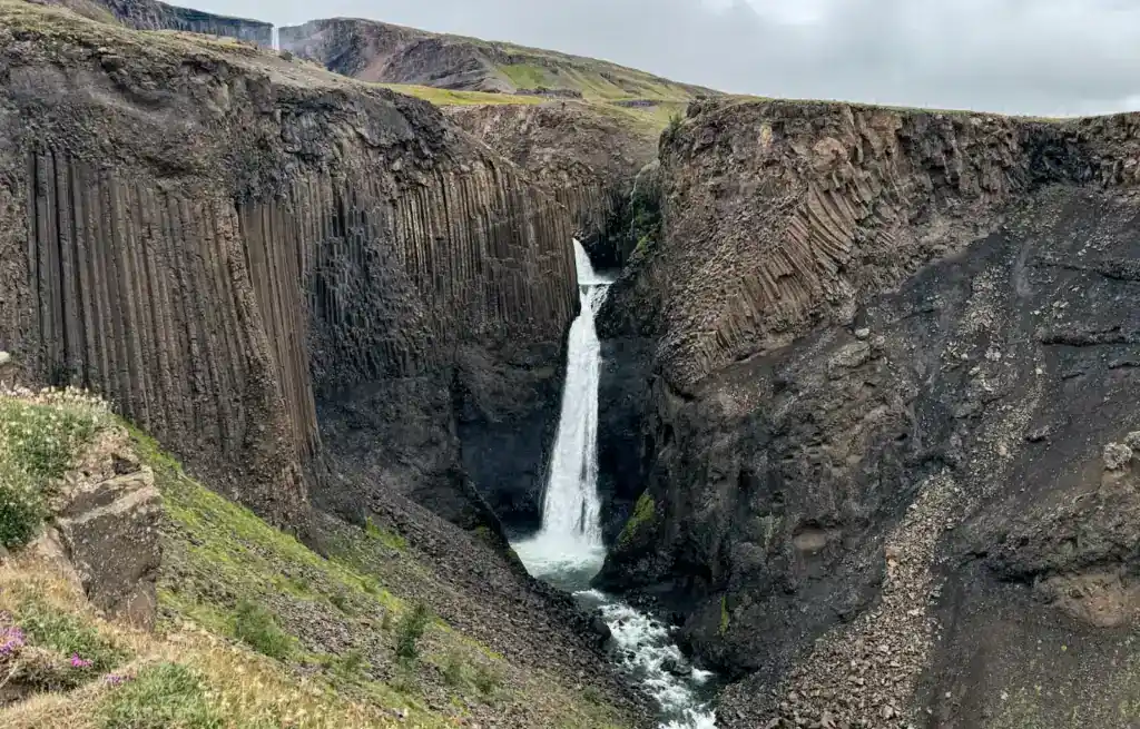 A tall waterfall cascading down a narrow, dark volcanic canyon with green vegetation on the edges.