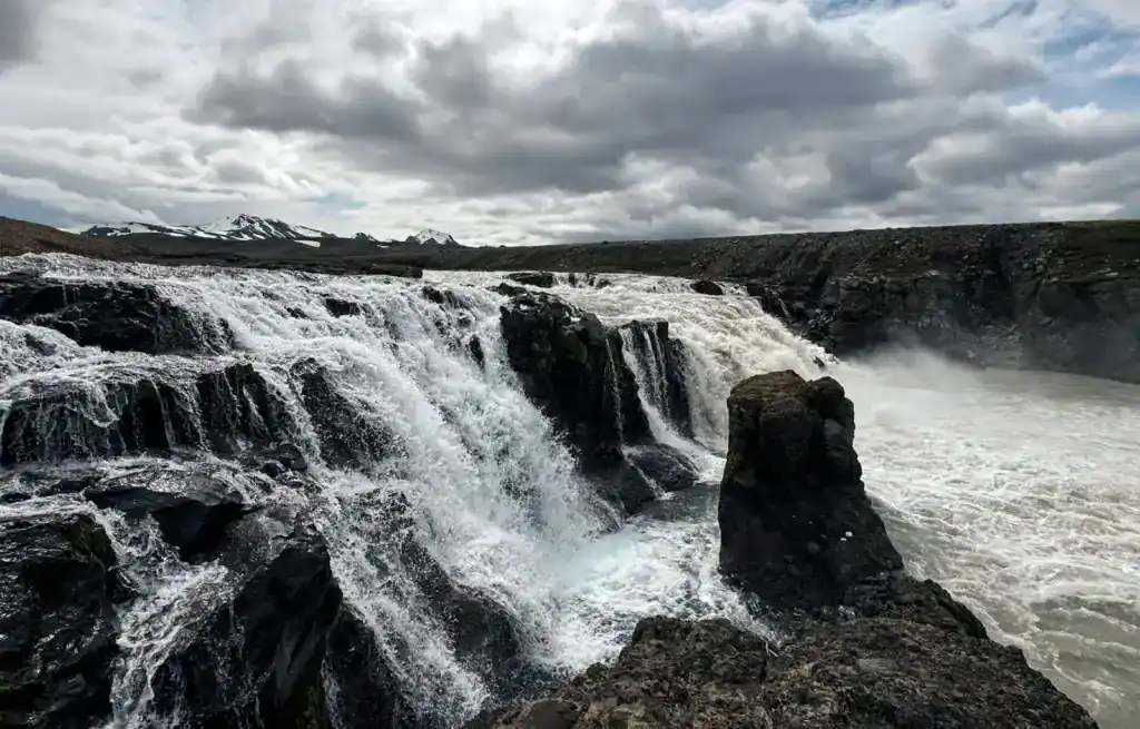 Powerful waterfall cascading over dark rocks under a cloudy sky in Iceland.