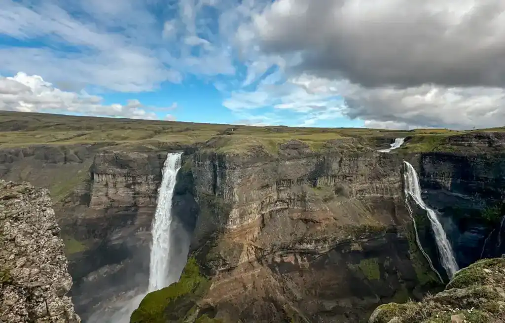 Two waterfalls cascade down steep cliffs under a partly cloudy sky in a rugged landscape.