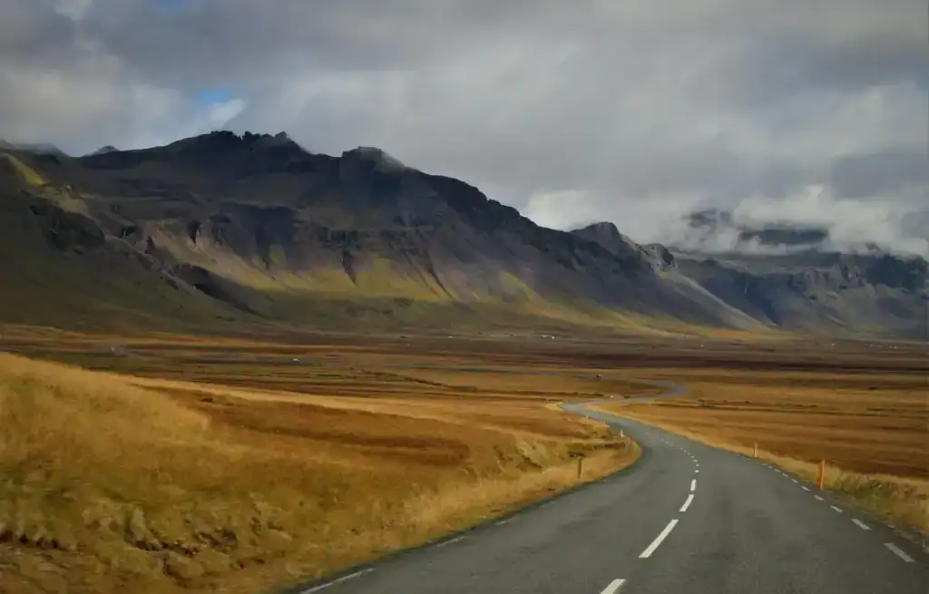 Winding road through vast Icelandic plains with mountains in the background under cloudy sky.