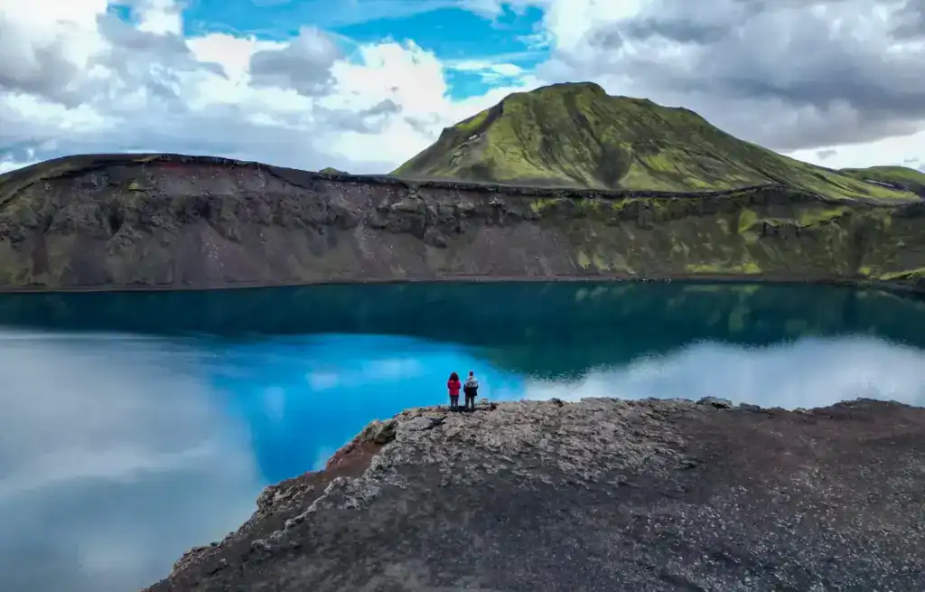 Two hikers stand on a rocky ledge overlooking a blue volcanic crater lake with green hills and cloudy sky.