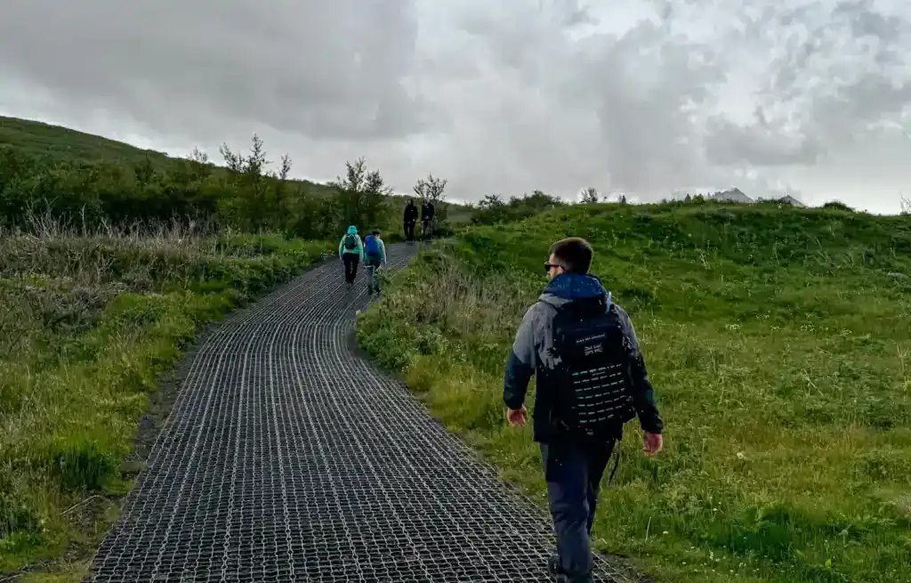 Hikers walking along a winding path through green hills under cloudy skies.