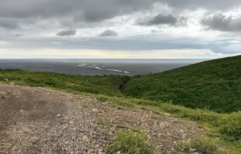 View over green hills towards a flat coastline with a river and cloudy sky.