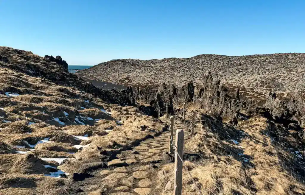 Stone pathway leading through rugged, grassy landscape towards distant cliffs and the sea under clear blue sky.