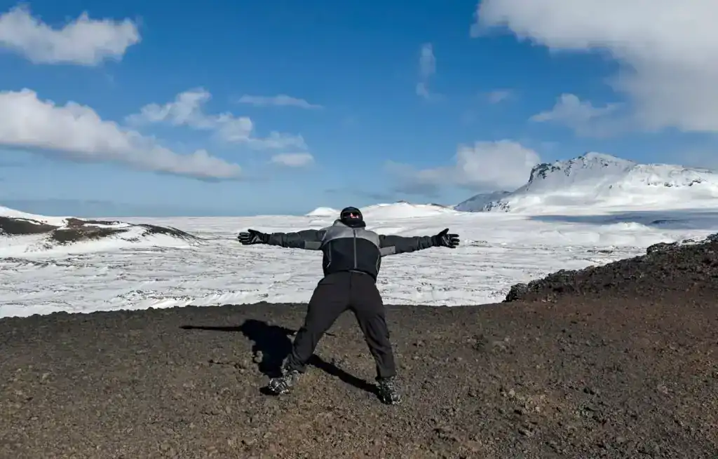 Person standing with arms outstretched on volcanic terrain with snow-covered mountains in the background.