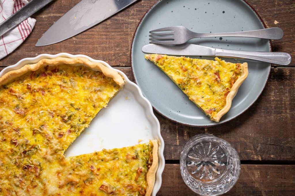 Ein angeschnittenes Stück Lauch Quiche auf einem Teller, daneben frischer Lauch und Speckwürfel als Zutaten für das Quiche Lorraine Rezept.