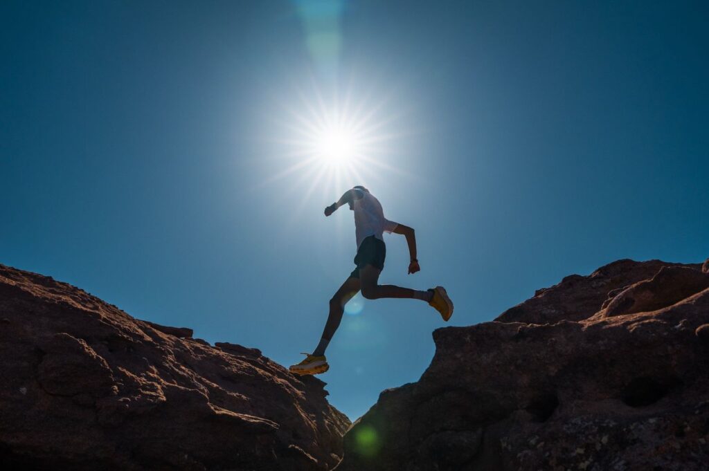 A photo of a man jumping across two ridges with the sun beaming down