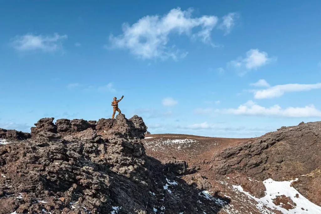 A hiker in a brown outfit stands triumphantly atop the rugged, dark volcanic rocks of Saxhóll Crater in Iceland. The landscape features jagged lava formations with patches of snow, set against a backdrop of rolling volcanic terrain and a bright blue sky with scattered clouds. The contrast between the rough, barren ground and the vast open sky creates a striking and adventurous scene.