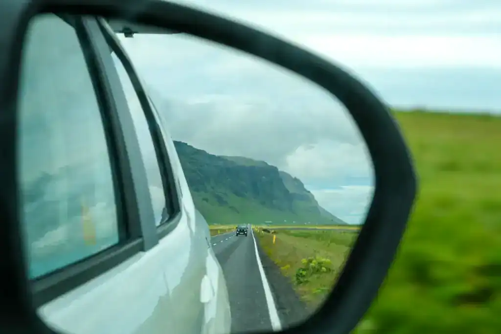 View through a car’s side mirror showing a winding coastal road in Iceland, lined with green fields and flanked by steep cliffs under a cloudy sky. A black SUV follows behind, adding a sense of motion and travel.