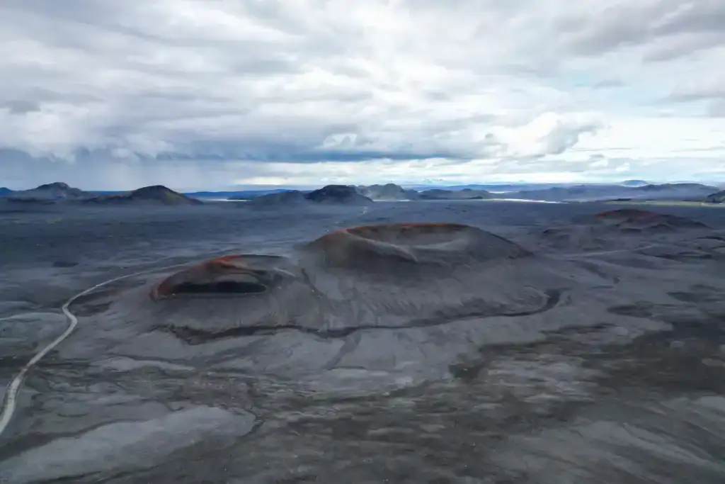 Aerial view of Iceland’s volcanic landscape featuring dark, ash-covered craters and barren plains. The terrain is marked by red-tinged ridges and winding paths, with distant mountains and a cloudy sky creating a dramatic, remote atmosphere.