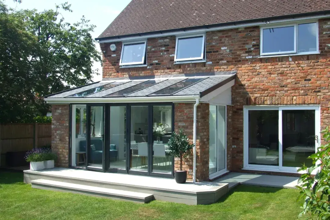 Lean-to conservatory with a low-pitched glazed roof, dark sliding doors and raised patio beside a red brick house.