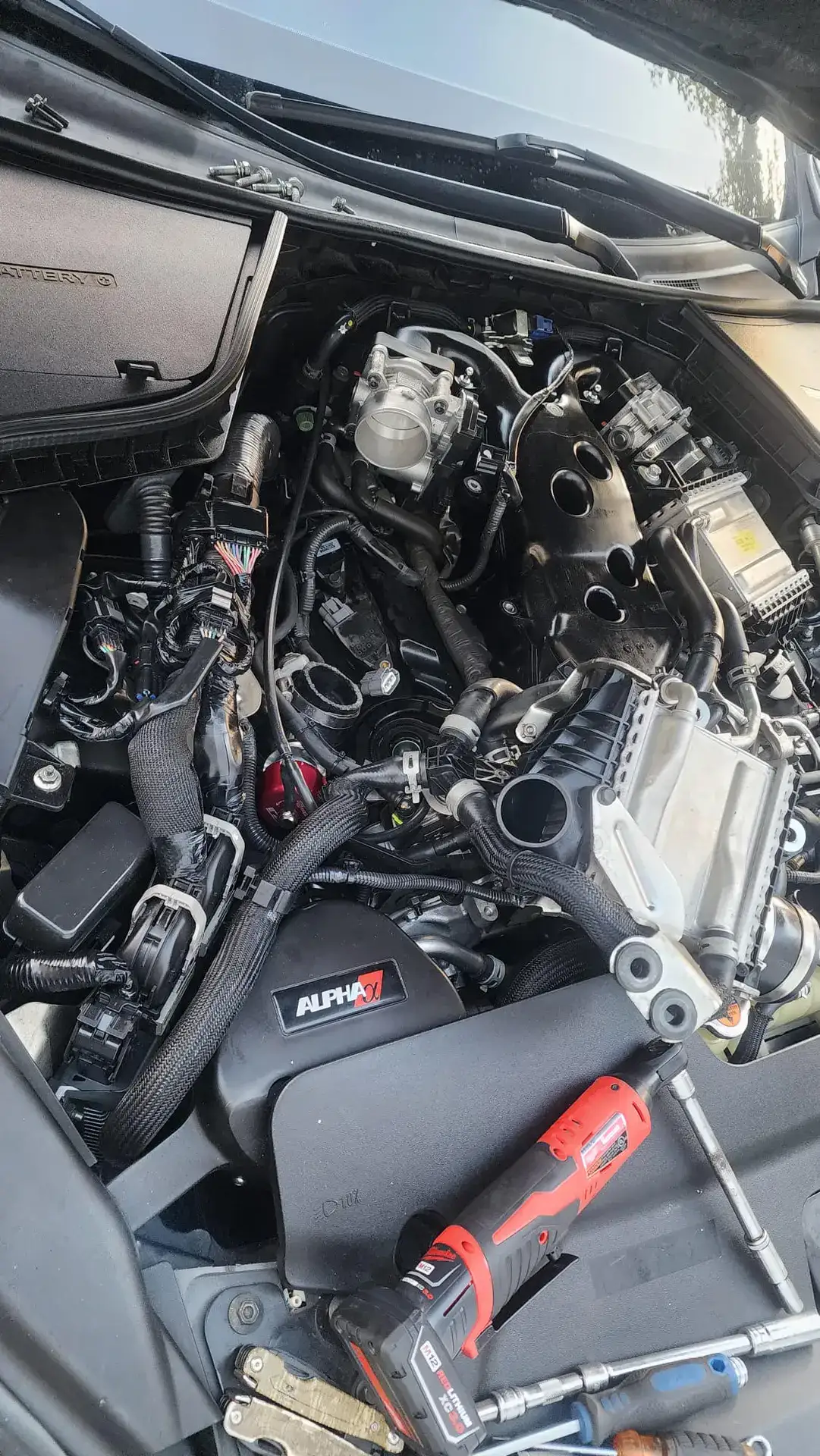 Car engine bay with the hood raised, exposing wiring, hoses, and metal engine components; a red and black drill rests on the nearby surface as tools are scattered nearby.