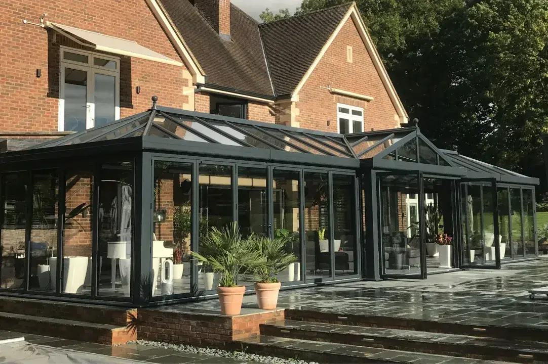 Large glass conservatory roof with dark frames, wide garden doors and black tiled patio outside a brick home.