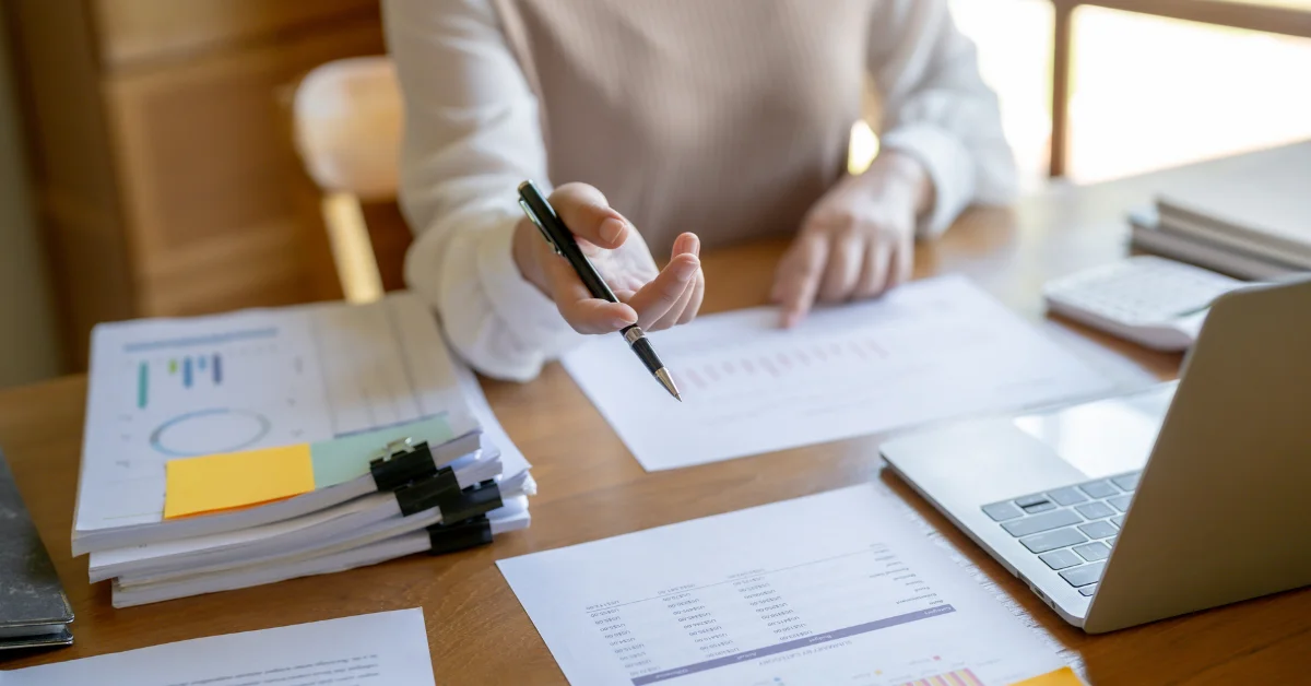 Image of a person holding a pen sitting an office table setting with papers and laptop | Advivo