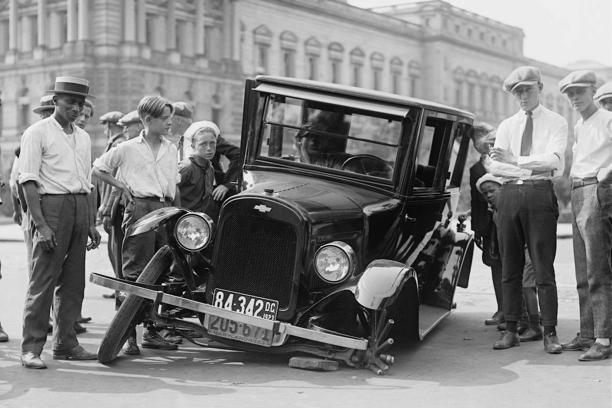 Schwarz-Weiß-Foto von ca. 1920, das eine Gruppe Männer und Jungen zeigt, die ein beschädigtes Auto mit eingeknicktem Vorderrad betrachten.