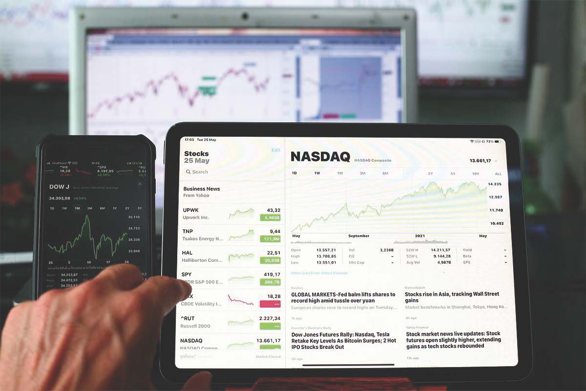 Hand zeigt auf Tablet mit NASDAQ-Chart; daneben Smartphone mit Dow Jones und Kursdaten.