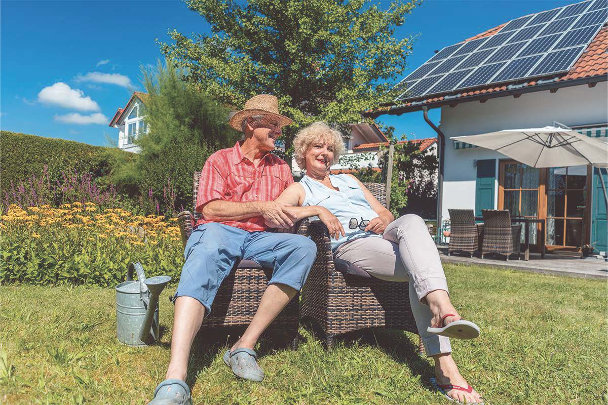 Seniorenpaar sitzt entspannt im Garten vor einem Haus mit Solaranlage bei sonnigem Wetter.