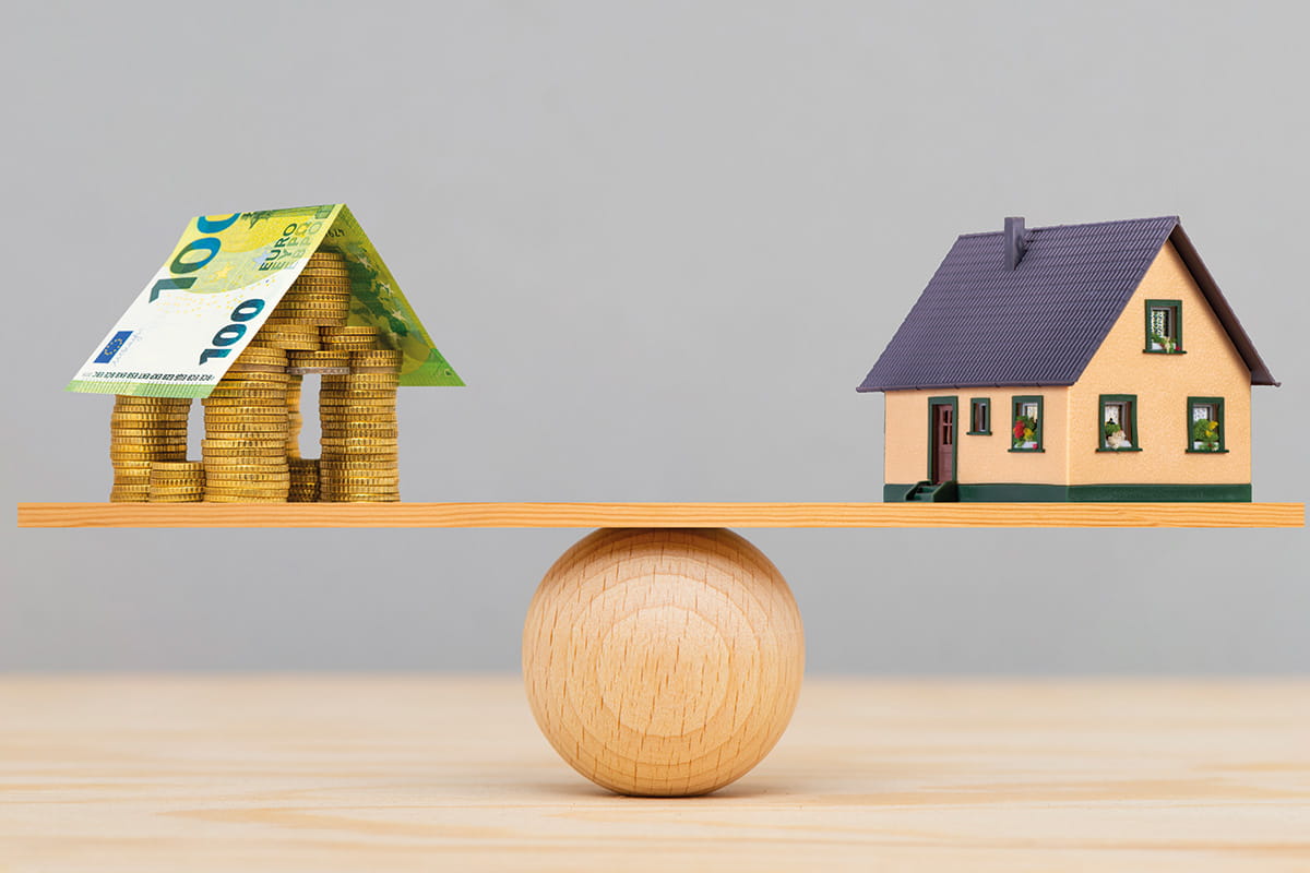 Haus aus Münzen und 100-Euro-Schein balanciert mit Modellhaus auf Holzbrett