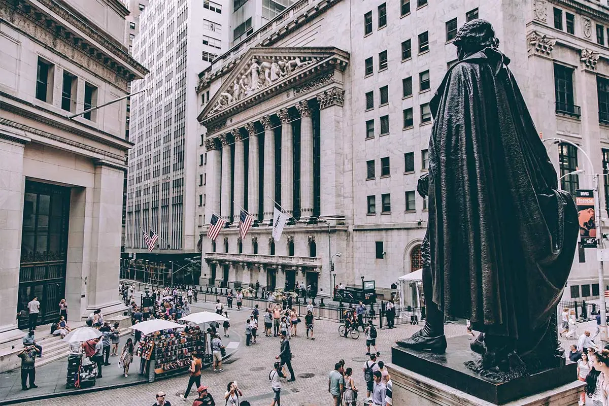 Blick auf die New Yorker Börse mit Flaggen, Passanten und Statue im Vordergrund.