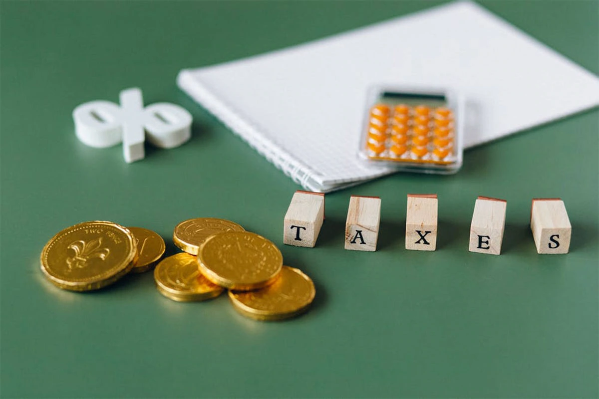 Goldmünzen, Holzwürfel mit dem Wort "TAXES", ein Block und ein Taschenrechner liegen auf einem grünen Tisch.