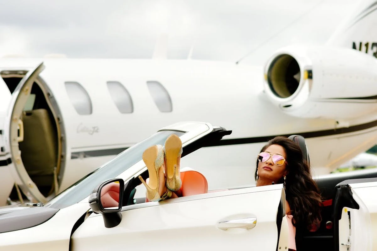 Frau in einem weißen Cabrio mit Füßen auf dem Armaturenbrett vor einem privaten Jet.