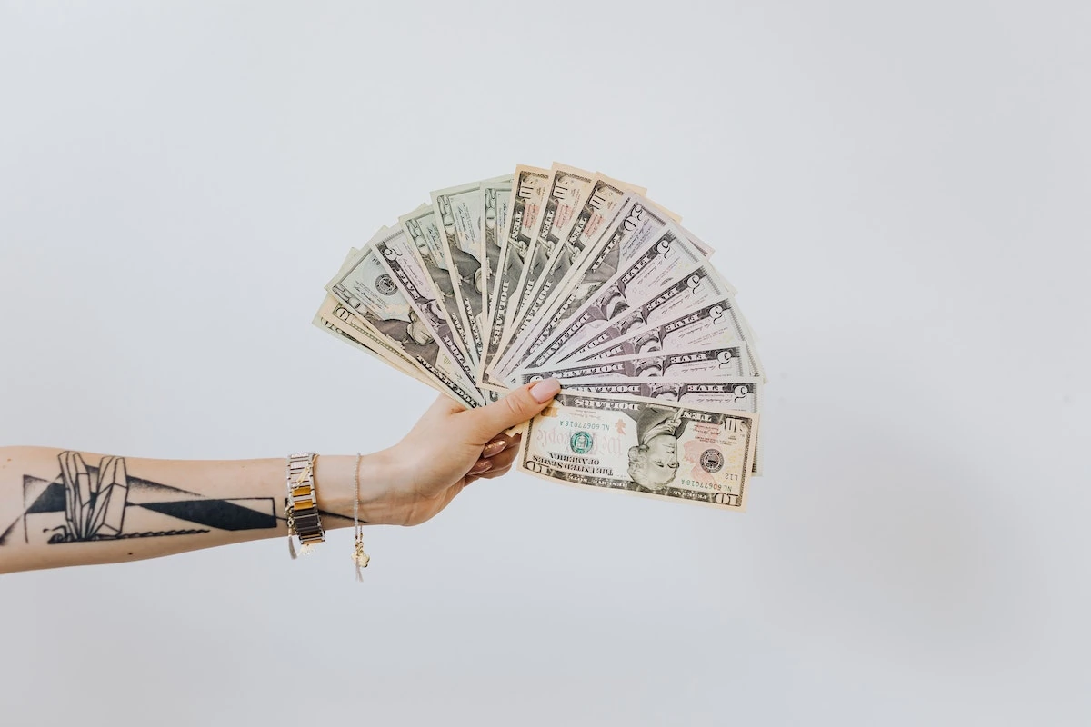 Eine tätowierte Hand hält eine Fächeranordnung von US-Dollar-Banknoten in verschiedenen Stückelungen vor einem weißen Hintergrund.