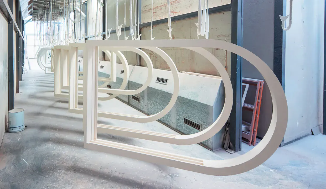 Industrial setting with multiple curved metal frames hanging in a row, likely being spray-painted or finished.