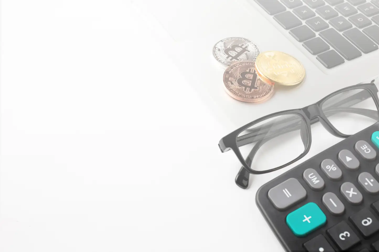 A pair of black glasses, a calculator, and three cryptocurrency coins (silver, copper, and gold) are placed on a laptop keyboard. The background is white, creating a clean and minimalistic setup.