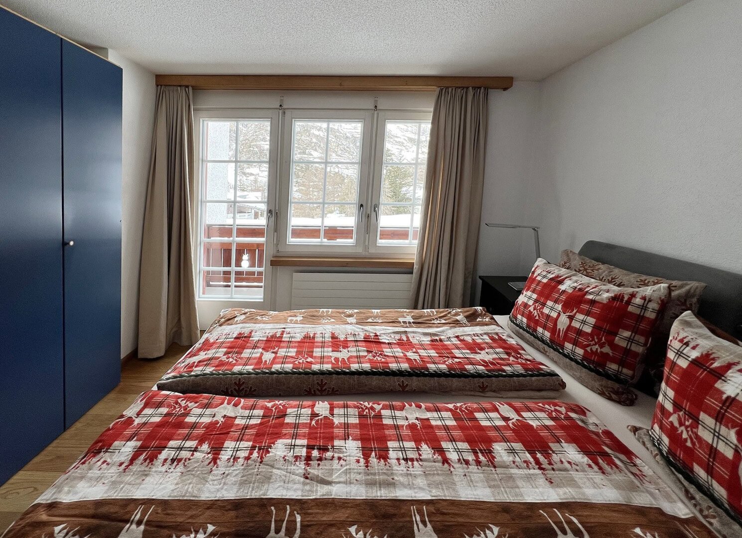 Gemütliches Schlafzimmer mit Doppelbett, rotem Karo-Bettwäsche, großen Fenstern und Blick auf die Natur, ideal für einen erholsamen Aufenthalt bei Matterhorn Home Apartments.