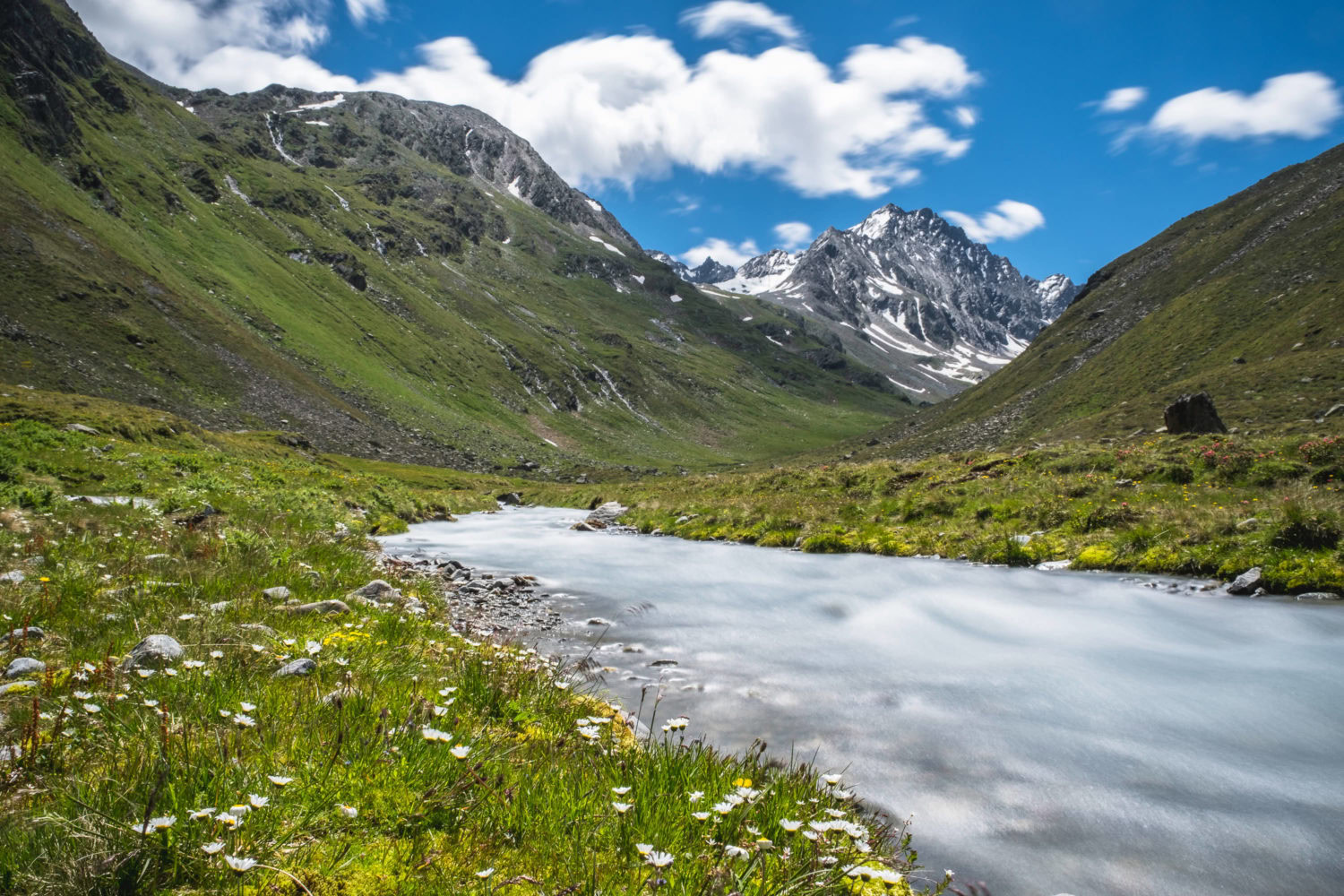 Fluss fließt durch Wiesen mit Berghängen