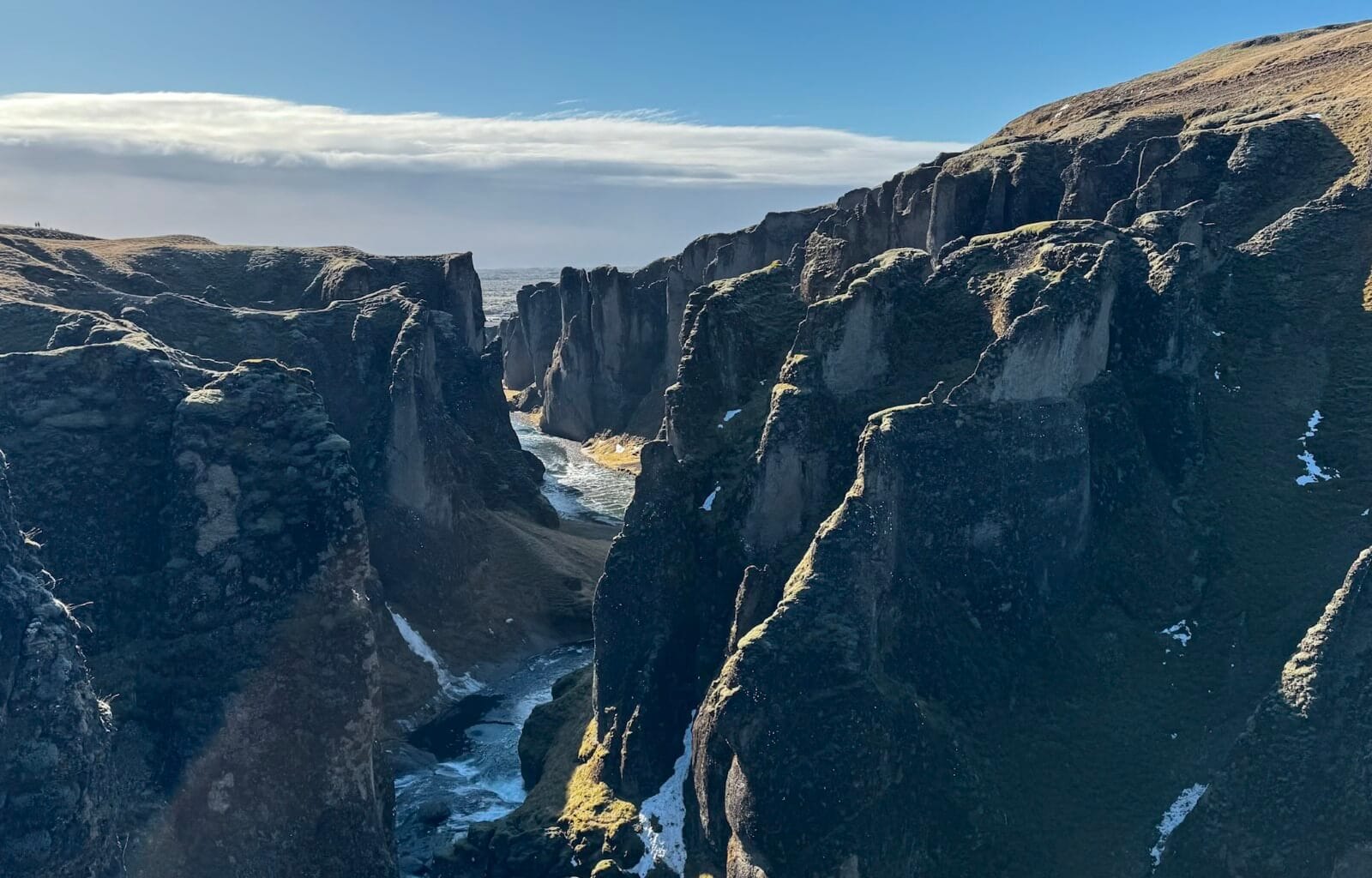 Fjaðrárgljúfur Canyon in Iceland stretches into the distance, its dramatic moss-covered cliffs rising steeply on either side of a winding river. The sunlight casts shadows on the rugged rock formations, highlighting the deep crevices and textures of the canyon walls. A bright blue sky and distant rolling hills complete this breathtaking natural landscape.