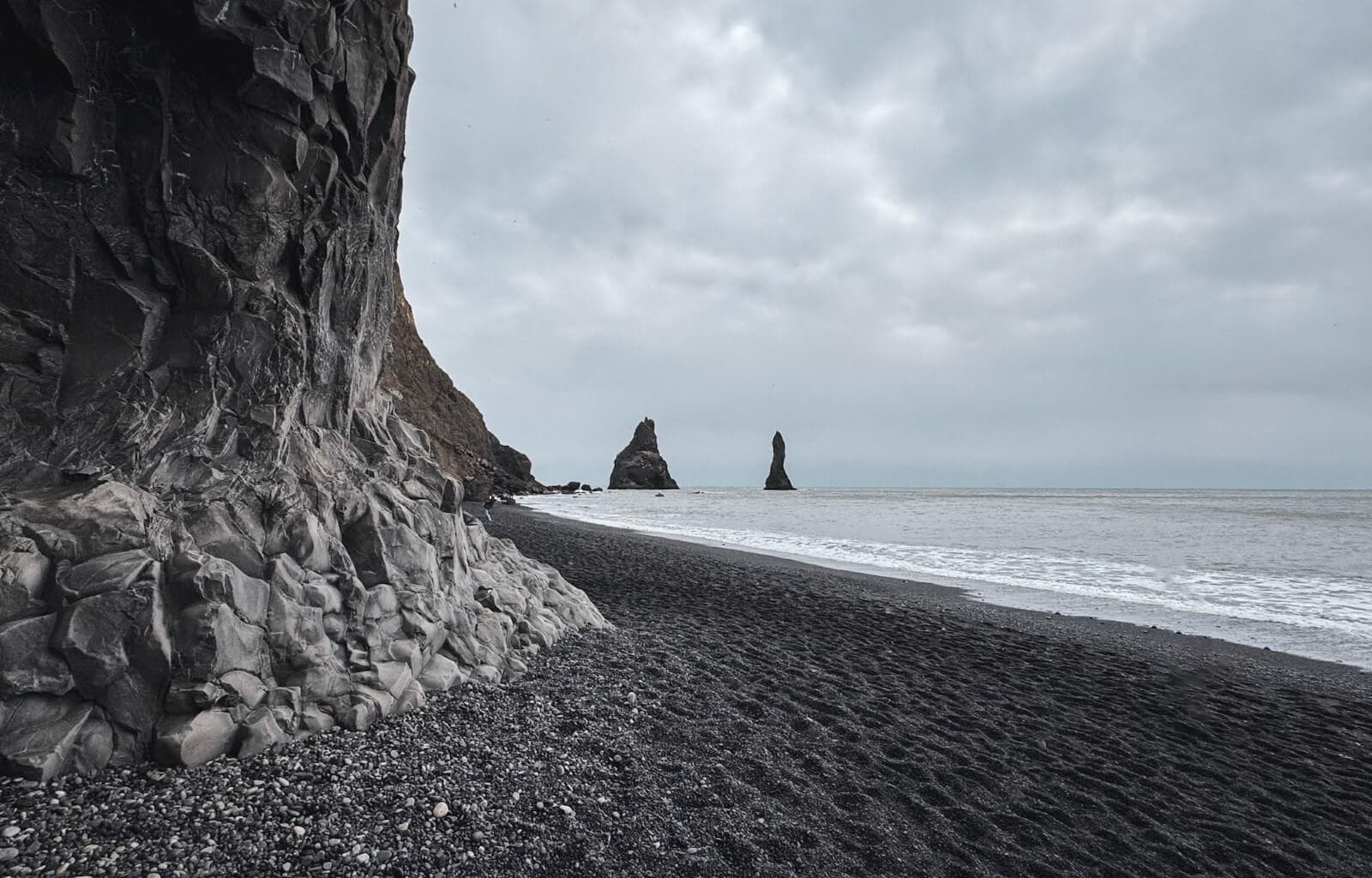 Dramatic basalt cliffs rise along the edge of Reynisfjara, Iceland’s famous black sand beach. The dark volcanic shoreline stretches toward the ocean, where powerful waves crash against the shore. In the distance, the Reynisdrangar sea stacks emerge from the misty waters, standing tall against the overcast sky. The moody atmosphere enhances the beach’s rugged and otherworldly beauty.