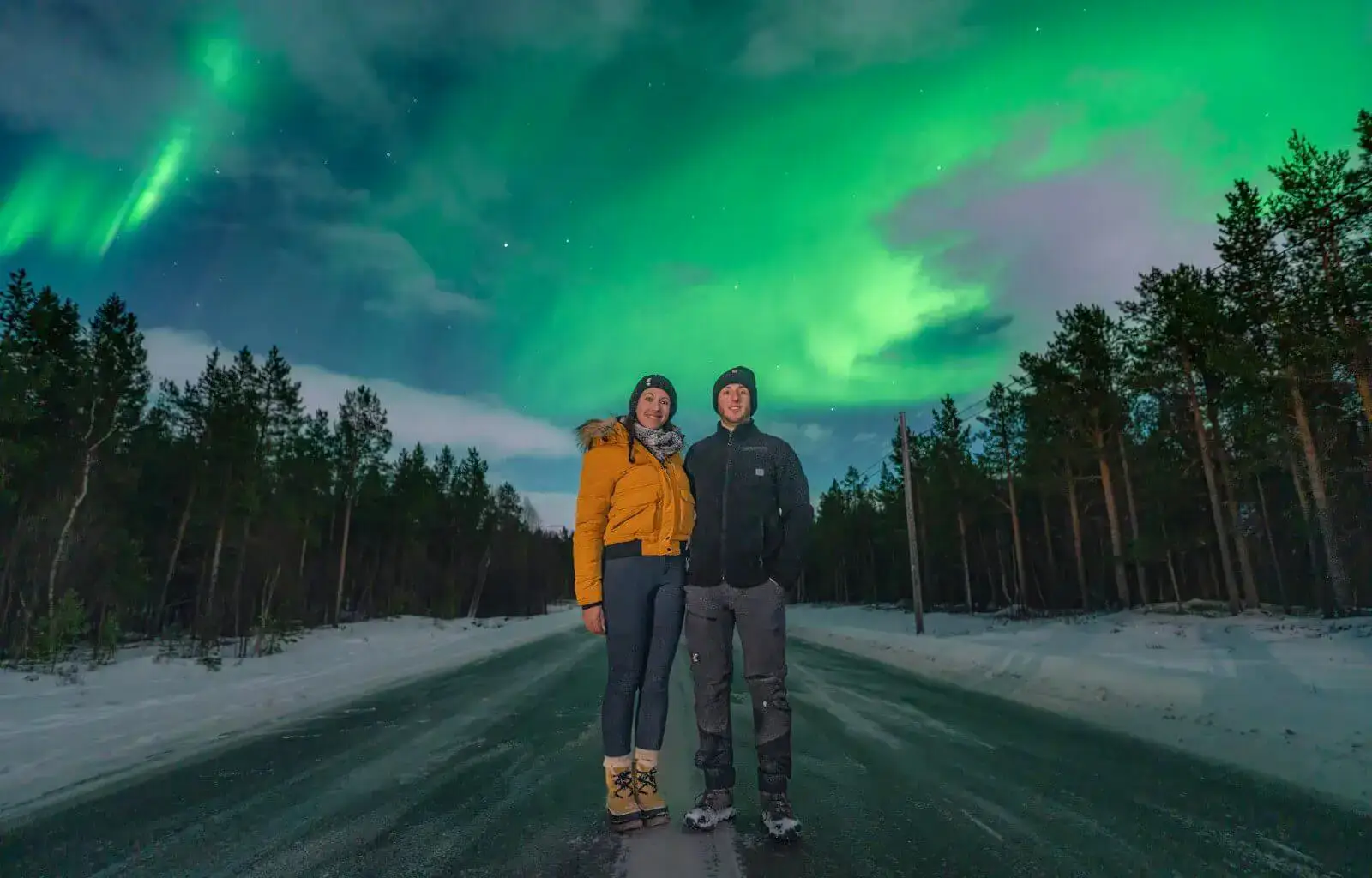 A couple stands on a snowy road at night, surrounded by a dense forest, as the vibrant green Northern Lights dance across the sky above them. The woman wears a yellow winter jacket, a beanie, and warm boots, while the man is dressed in dark outdoor clothing. Both are smiling, enjoying the breathtaking Arctic scenery.
