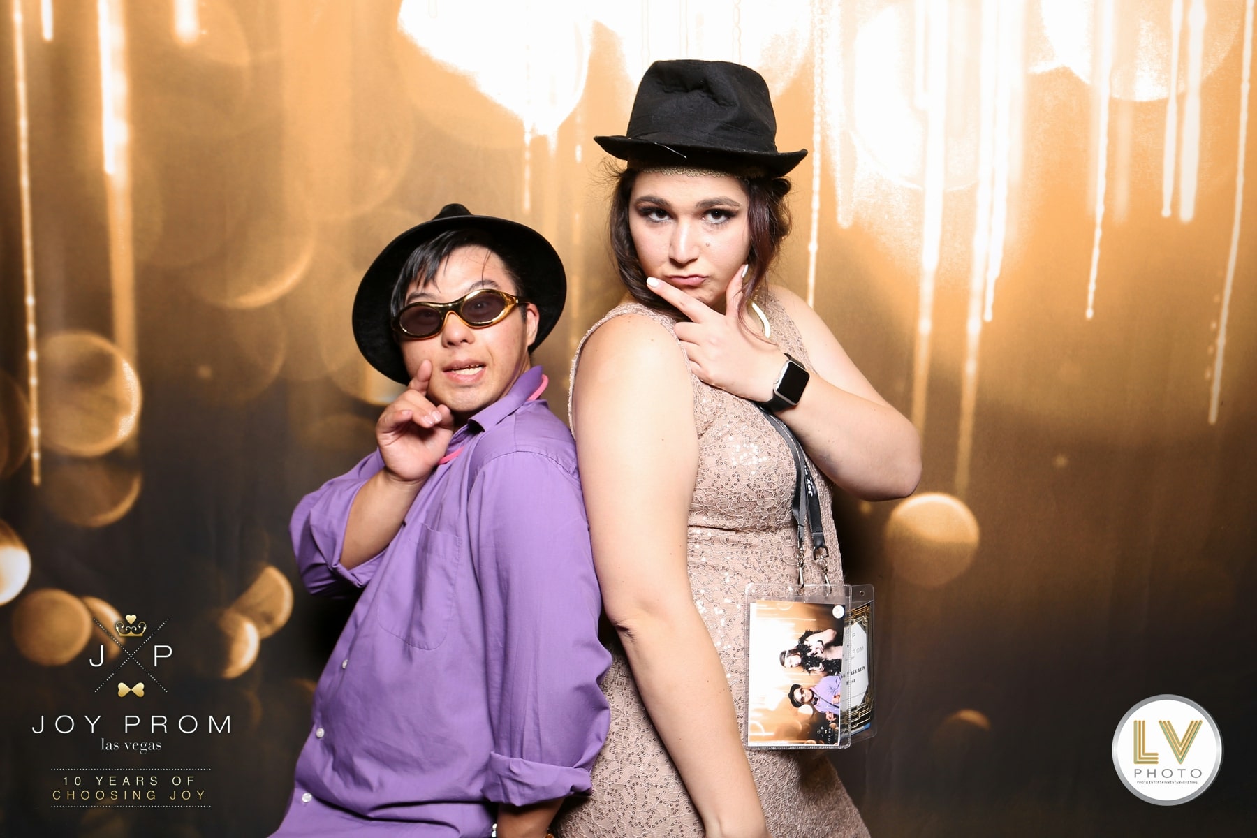 Two women posing in a photo booth with hats and sunglasses against a gold backdrop.