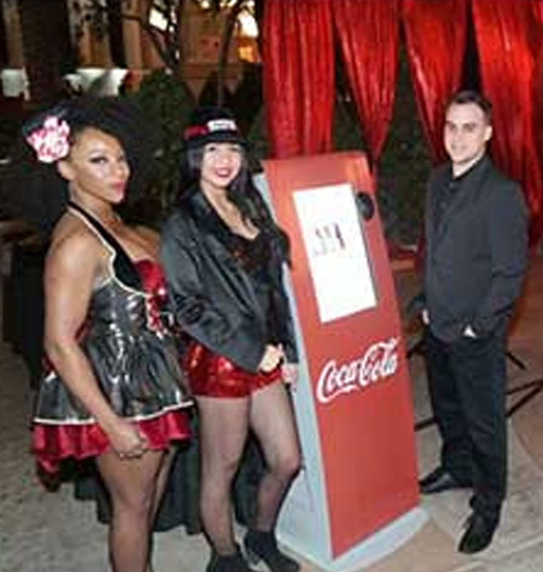 Three costumed people pose beside a Coca-Cola display and a red promo kiosk at an event.