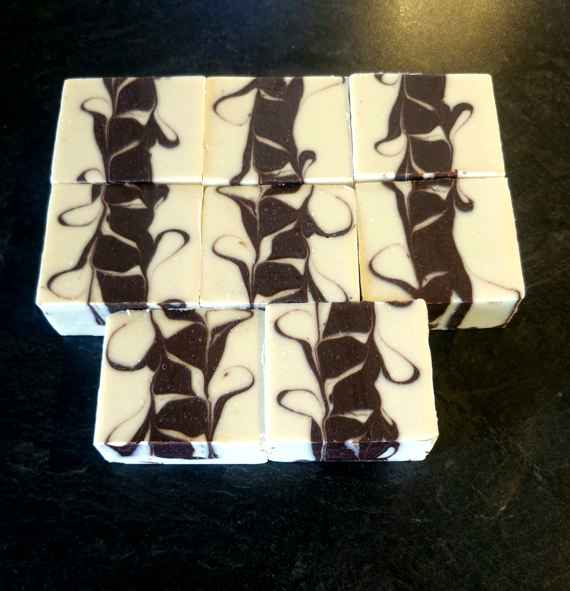 Nine bars of handcrafted grass-fed tallow soap in a rich dark chocolate brown and cream swirl pattern, coloured with cacao and coffee, arranged on a dark slate background, handmade by Fatglow in Taranaki New Zealand