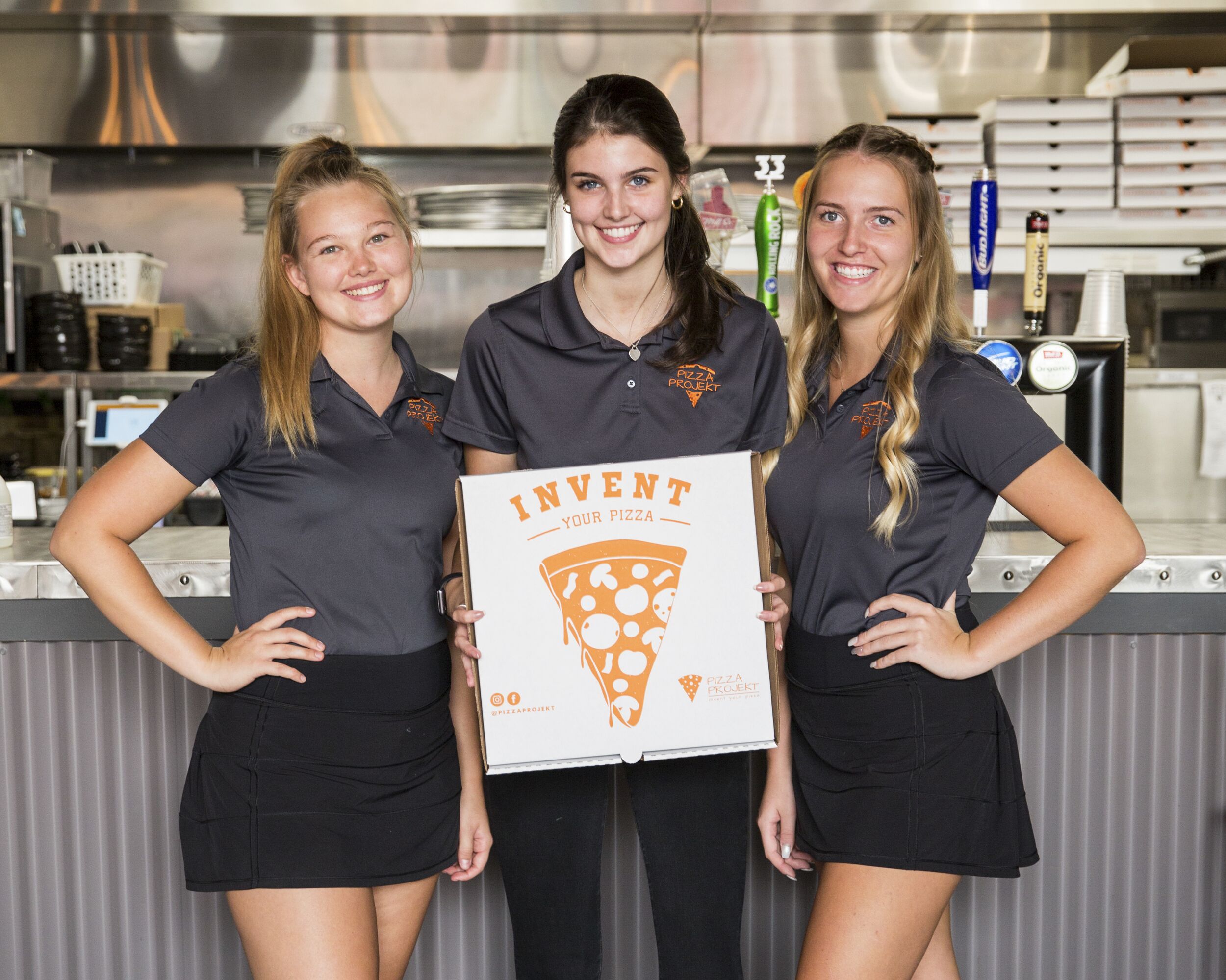 3 pizza projekt servers smiling and holding up a pizza box infront of the bar area.