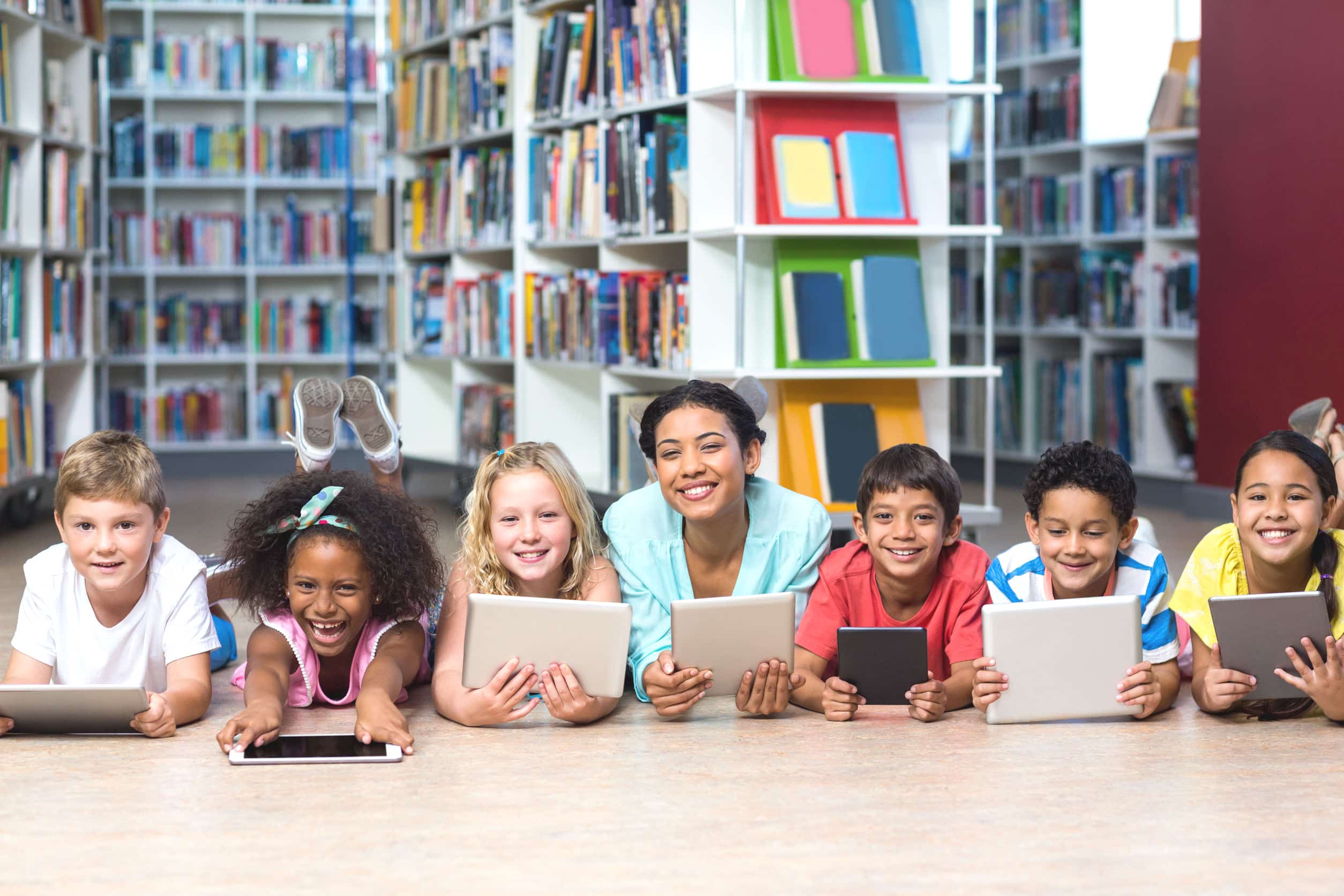 Portrait of smiling teacher with students using digital tablets while lying down in library