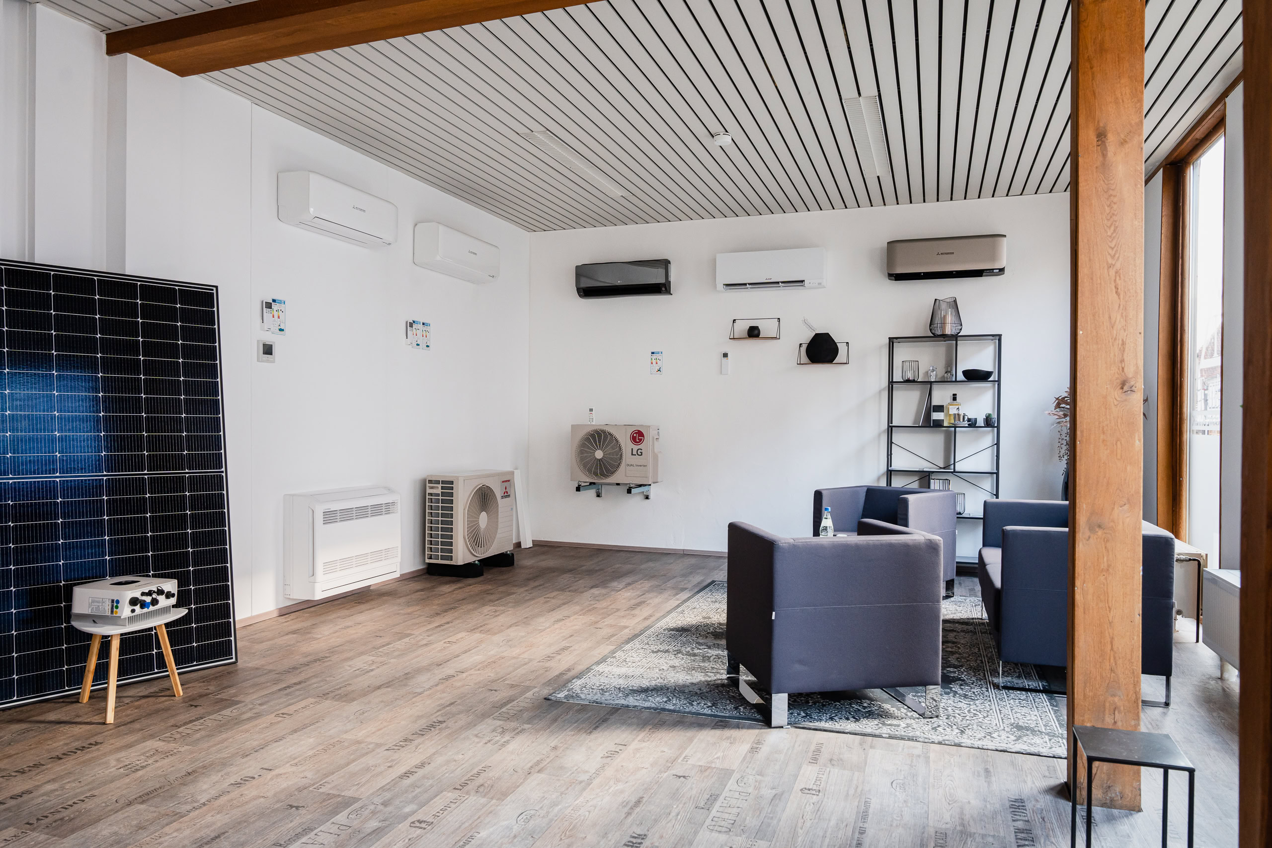 Über uns – Verkaufsraum von Delta-T Klimatechnik. Das Bild zeigt einen hellen Verkaufsraum von Delta-T Klimatechnik mit zwei Sesseln, einem Sofa, und diversen Ausstellungsstücken wie einem PV-Panel, mehreren Klimaanlagen-Innen- und -Außengeräten.