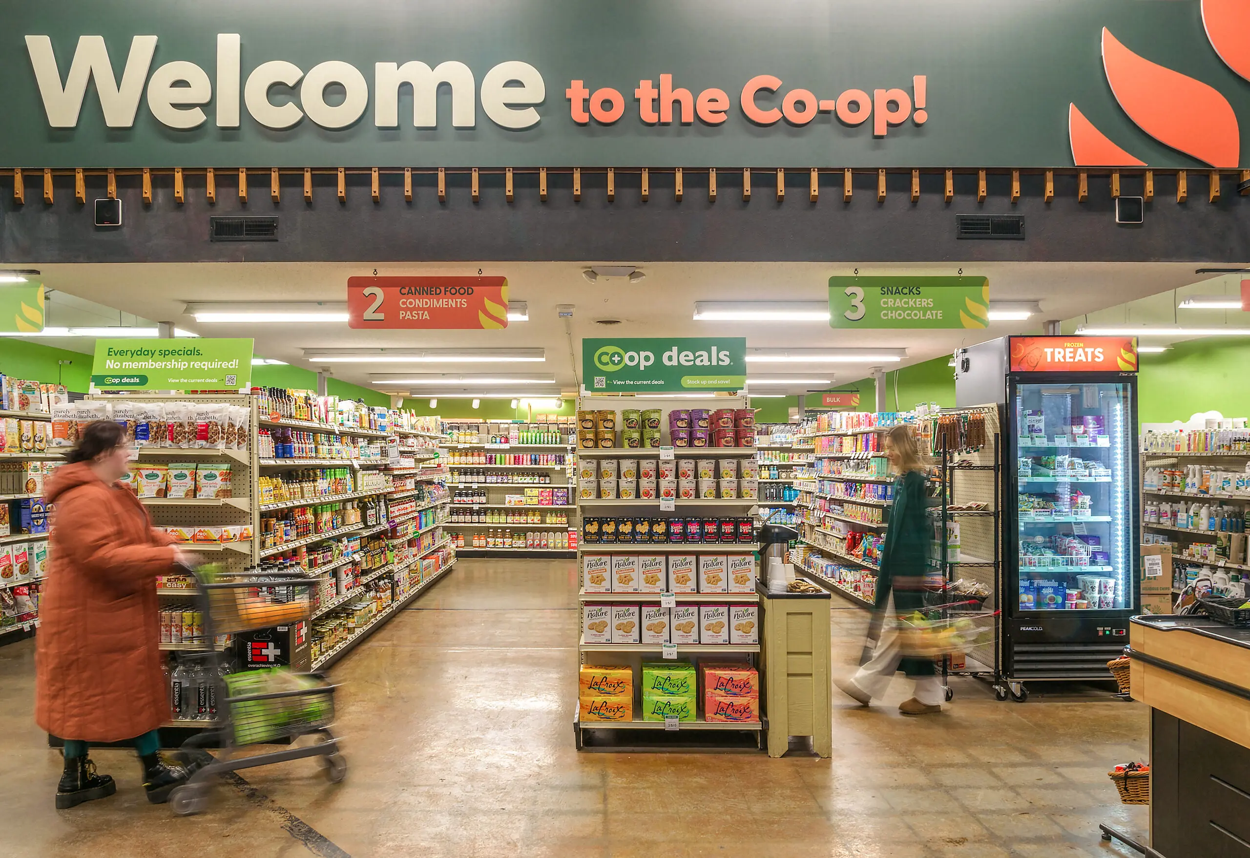 A welcoming entrance to a community-owned grocery co-op, featuring aisles stocked with organic, local, and natural foods. Shoppers browse fresh and sustainably sourced products, with prominent signage for co-op deals and everyday specials.